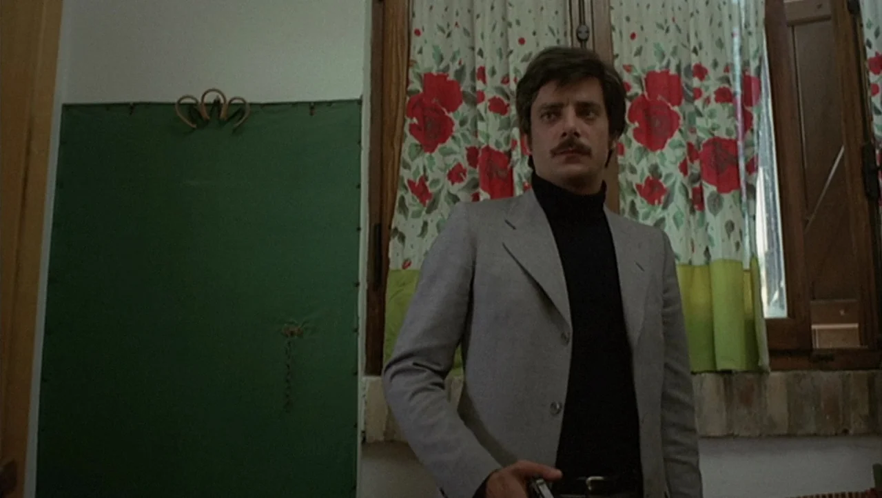 The Black Belly of the Tarantula (1971) movie still: medium — A man stands in an indoor space with floral curtains in the background. The man holds…