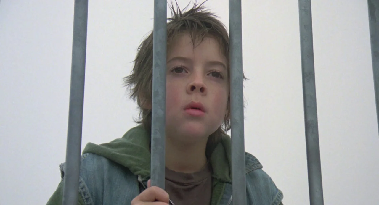 River’s Edge (1986) movie still: closeup — A young child stands behind a series of vertical grey metal bars. The background is a bright, washed-out…