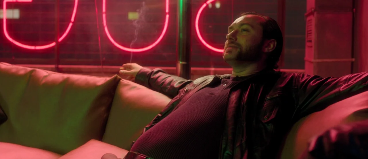 Dom Hemingway (2013) movie still: medium — A dimly lit lounge space features bright red neon signage on a window wall. A man sits comfortably on a…