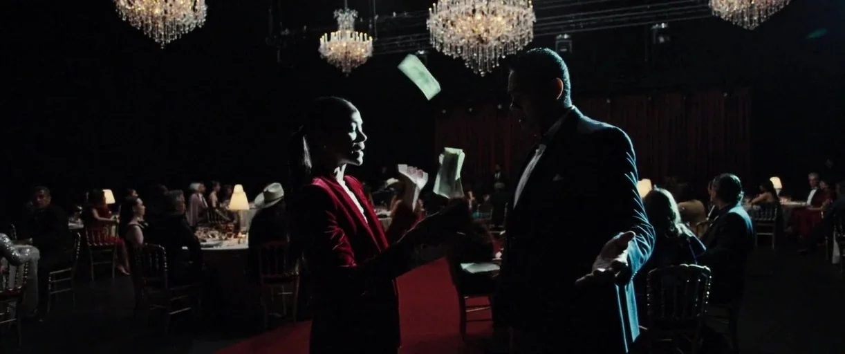 Emilia Pérez (2024) movie still: medium — A dark ballroom with crystal chandeliers illuminating a crowd seated at round dining tables. A woman in a…