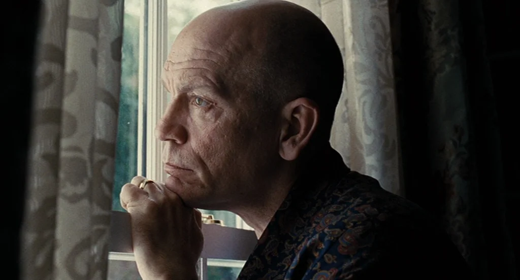 Burn After Reading (2008) movie still: closeup — A man looks through a window pane while standing beside white curtains. He rests his chin on his…