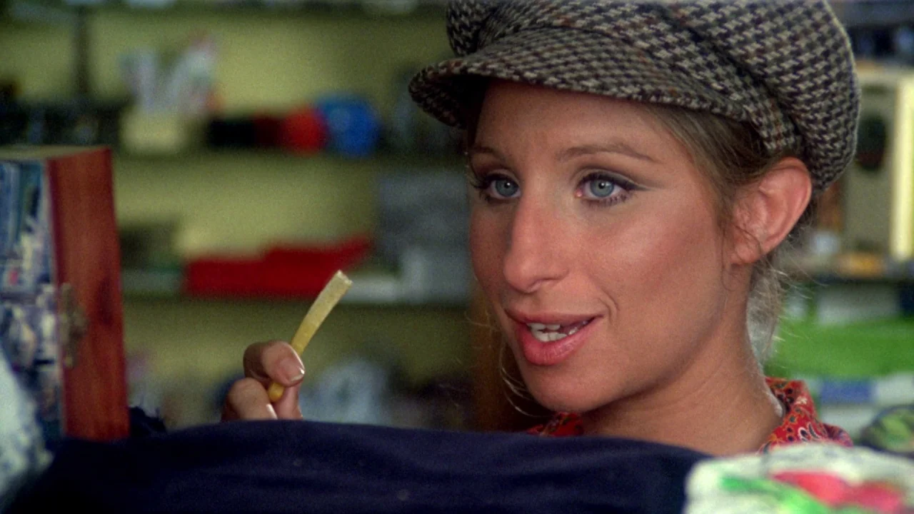What’s Up, Doc? (1972) movie still: closeup — A woman stands in a shop with shelves visible in the blurred background. She holds a single french fry…