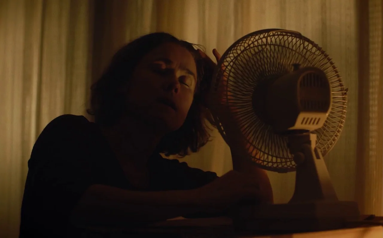 Knocking (2021) movie still: medium — A dimly lit room with draped curtains visible in the background. A woman leans over a small desk, positioned…
