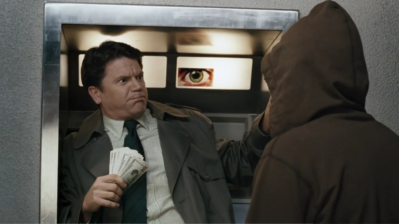 Fun with Dick and Jane (2005) movie still: medium — A man stands inside a narrow metal enclosure holding paper currency while facing a hooded…