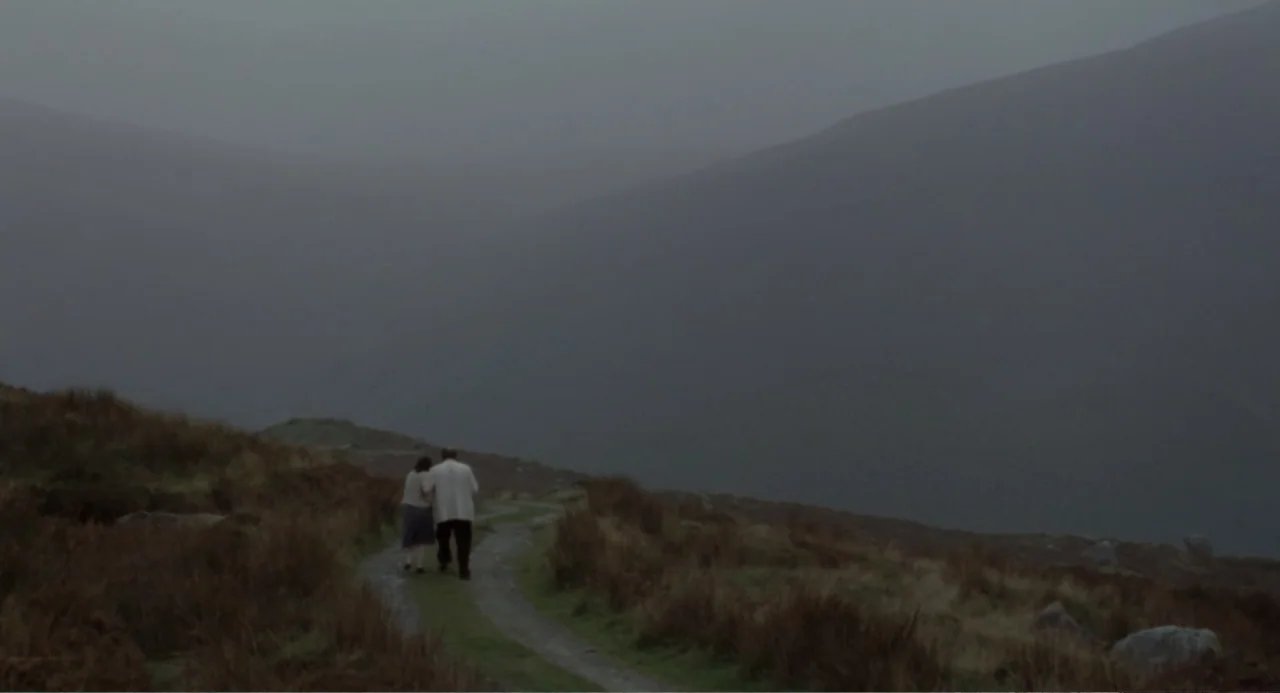 Dancing at Lughnasa (1998) movie still: wide — A desolate mountain valley under heavy cloud cover features a dirt path. Two figures walk away from…