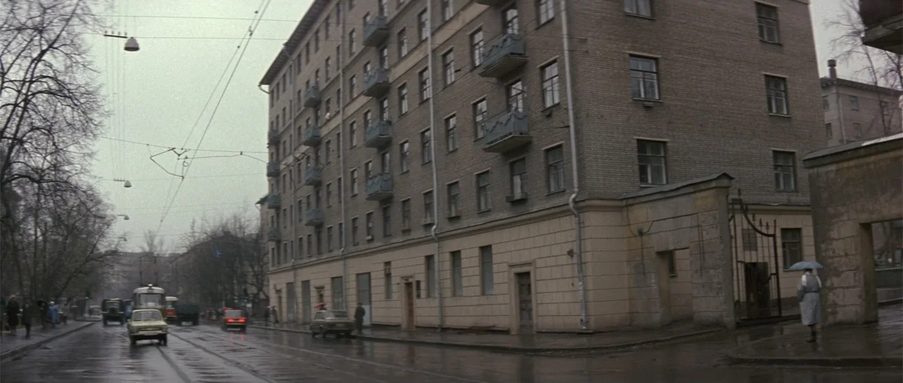 The Russia House (1990) movie still: establishing — A wet asphalt city street runs alongside a large, multi-story grey brick apartment building.…