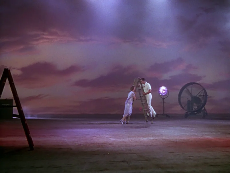 Singin’ in the Rain (1952) movie still: wide — A spacious soundstage features a large painted backdrop of purple and pink sunset clouds. Two…