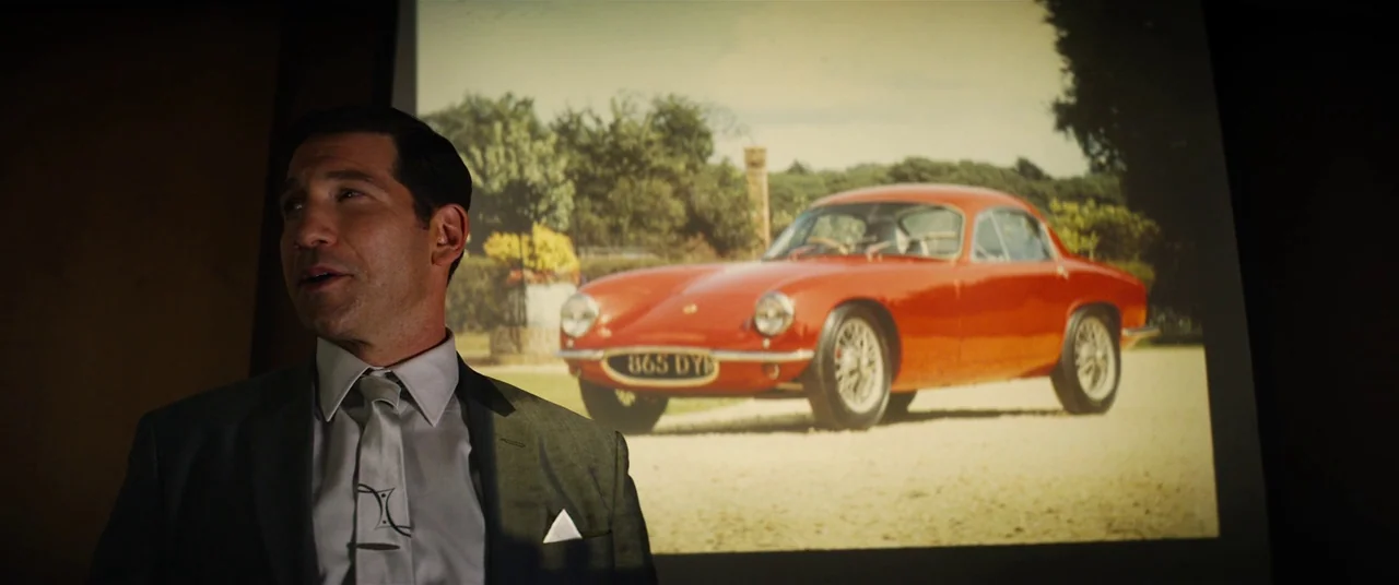 Ford V Ferrari (2019) movie still: medium — A dark, dimly lit conference space contains a projection of a red sports car. A man in a formal suit…