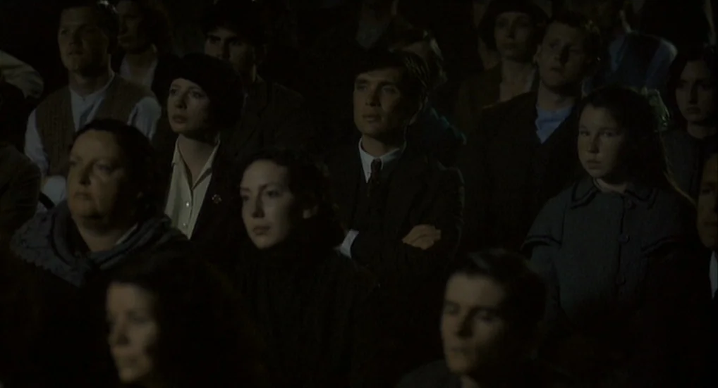The Wind That Shakes the Barley (2006) movie still: medium — A dimly lit theater audience sits in rows of seats, facing forward in silence. A man…