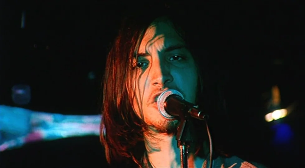 Scarlet Diva (2000) movie still: closeup — A dark concert venue with minimal light and obscured background elements. A man with long dark hair sings…