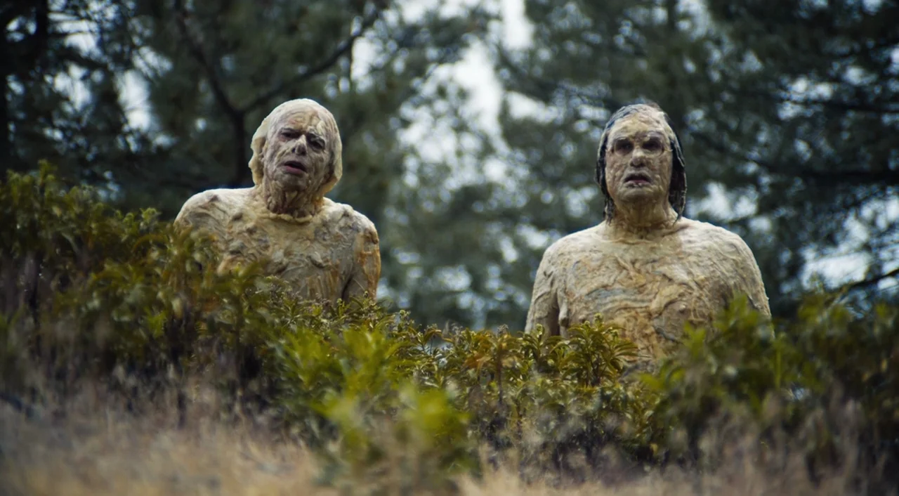 The Greasy Strangler (2016) movie still: medium — Two men covered in a thick, textured, flesh-toned substance stand within tall field grass. Behind…