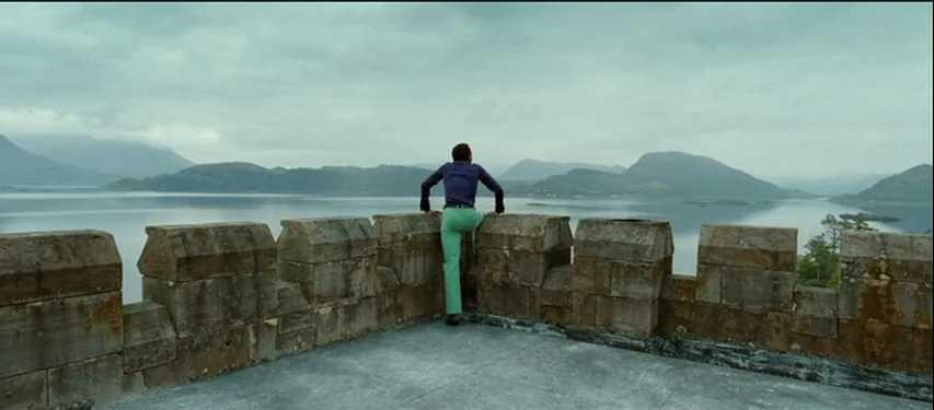 Mister Lonely (2007) movie still: wide — An outdoor stone wall overlooks a vast lake surrounded by mountains under overcast skies. A man stands on…