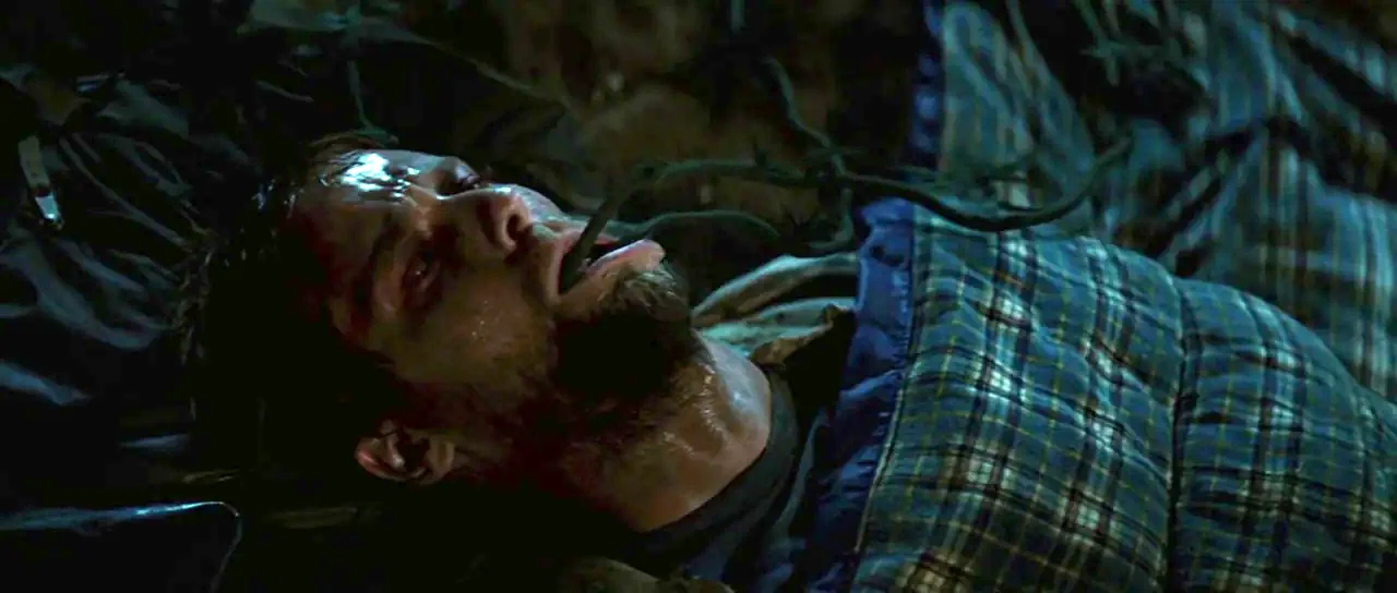 The Ruins (2008) movie still: closeup — A man lies within a patterned sleeping bag in a dark outdoor environment. A dark, thin vine or plant tendril…