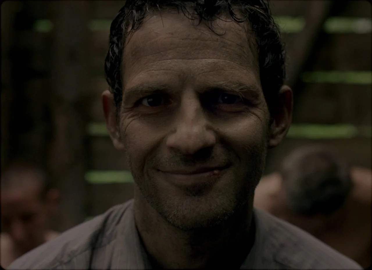 Son of Saul (2015) movie still: closeup — A dimly lit interior with horizontal wooden planks making up the walls. A man stares forward in a tight…