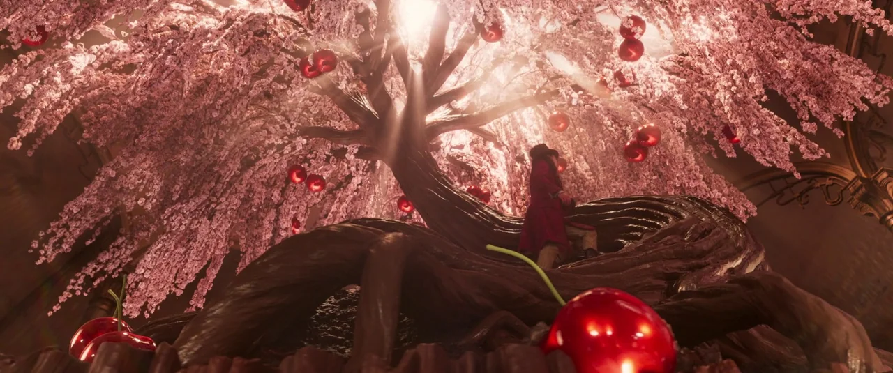 Wonka (2023) movie still: wide — A surreal room features a massive tree with chocolate-like bark and pink blossom branches. A man in a red coat sits…