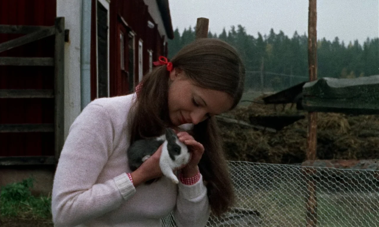 Thriller: A Cruel Picture (1973) movie still: medium — A teenage girl stands outdoors near a red wooden building and a wire fence. She holds a small…