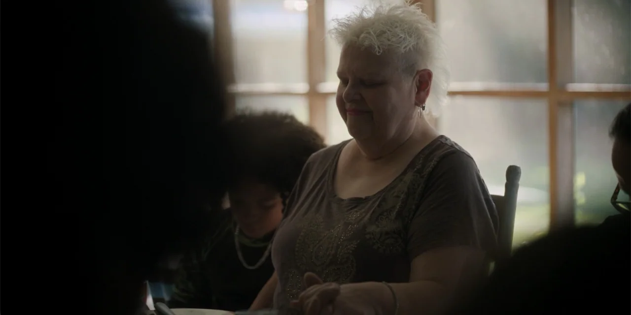Caddo Lake (2024) movie still: medium — A dimly lit room with a wooden window frame visible in the background. An older woman with short curly white…