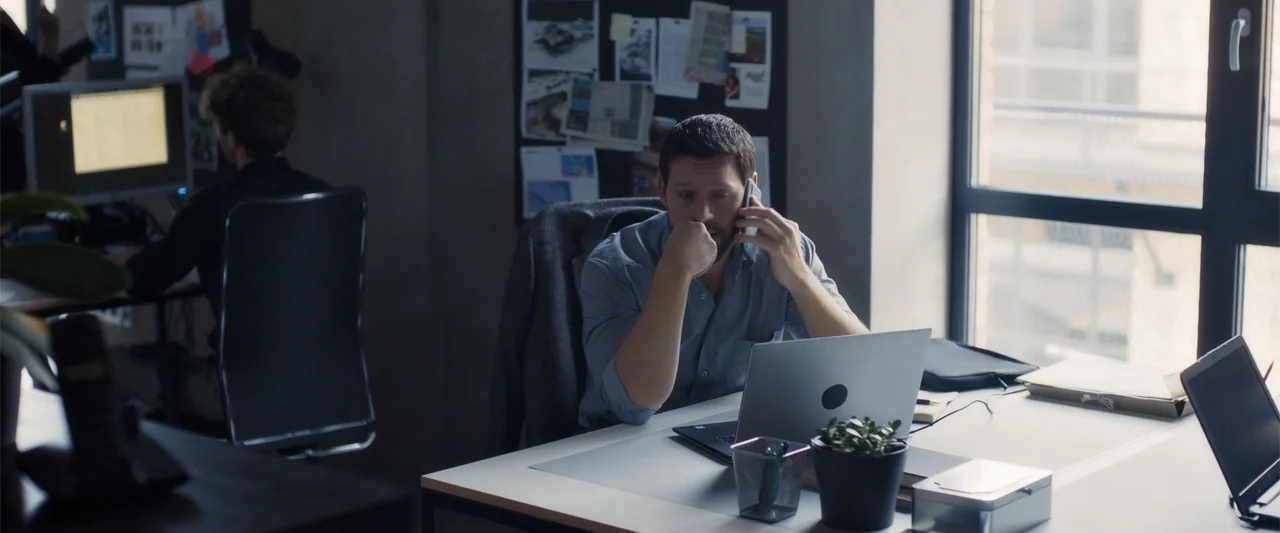 The Good Neighbor (2022) movie still: medium — An open plan office features desks with laptops and a window with natural light. A man sits at his…