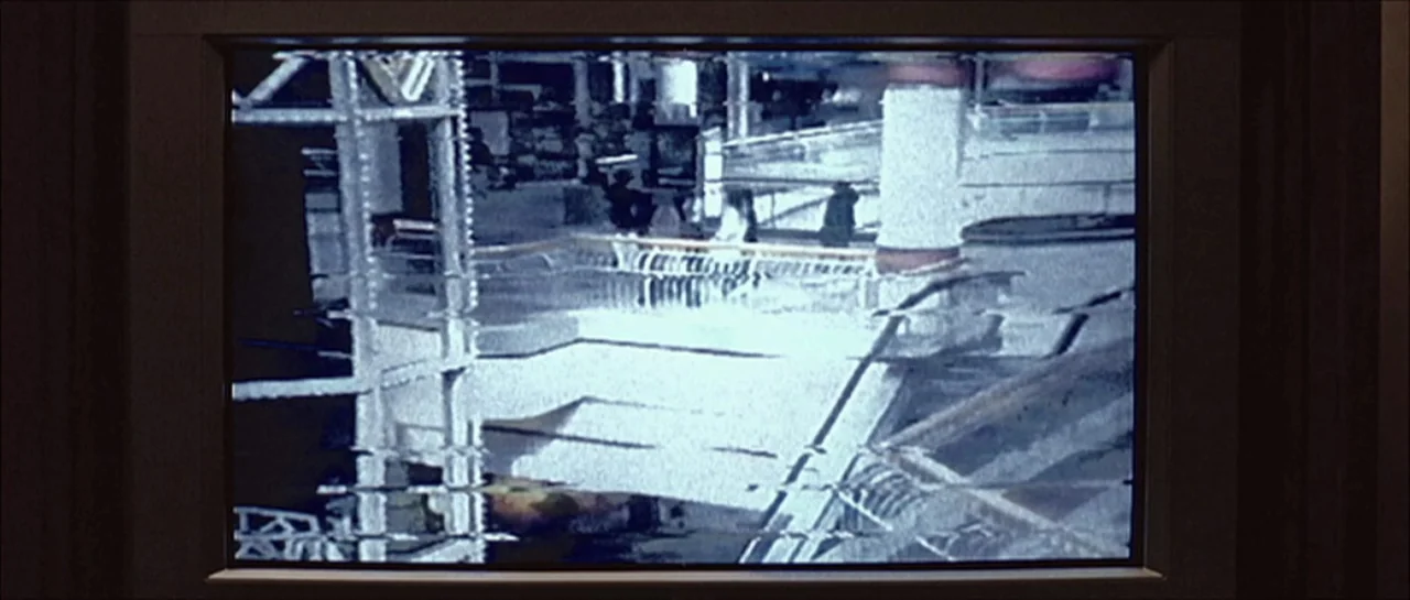 Fear X (2003) movie still: establishing — A high-angle security camera feed shows a multistory mall atrium with white floors. The image contains…