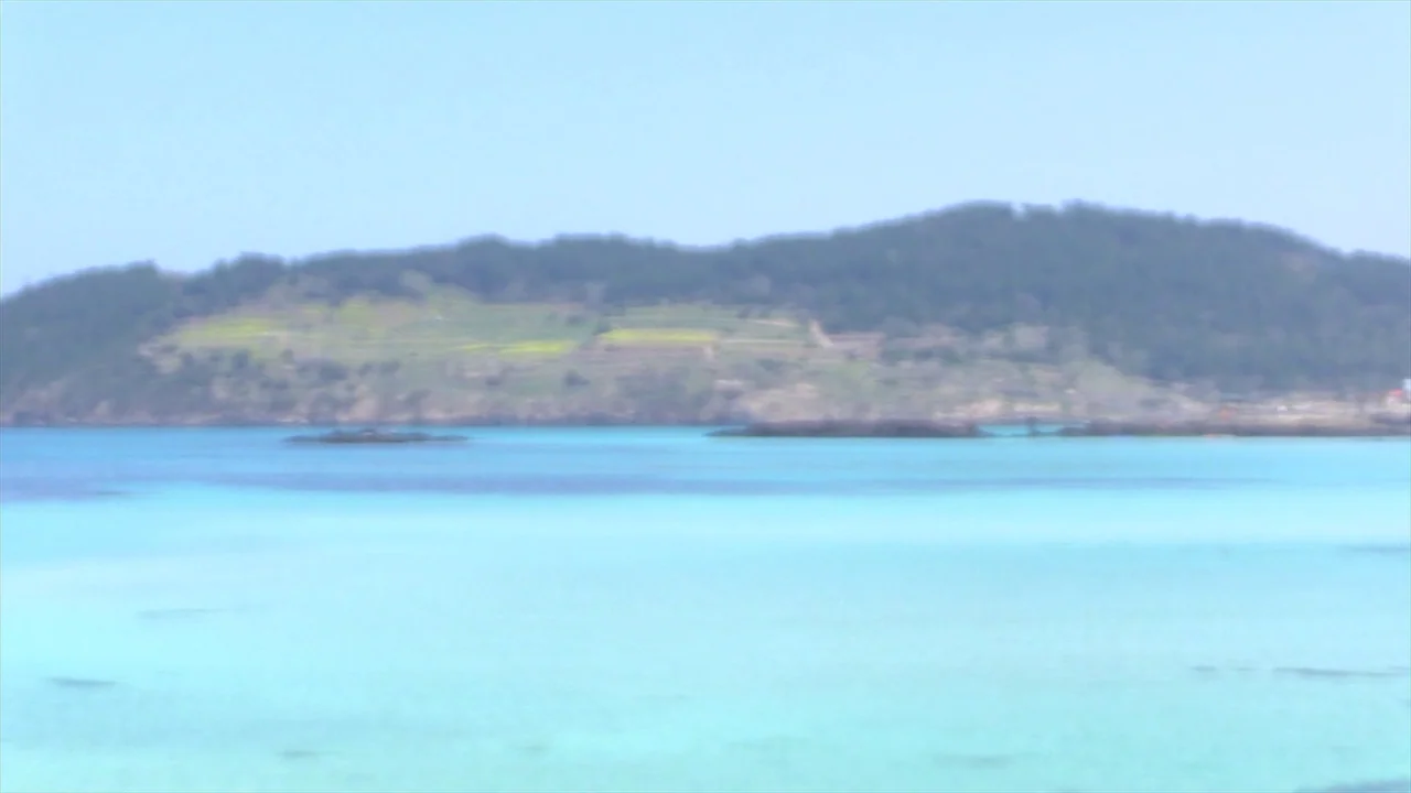 In Water (2023) movie still: establishing — A turquoise bay sits before a distant, hazy green hill under a clear sky. Small rocky islets break the…
