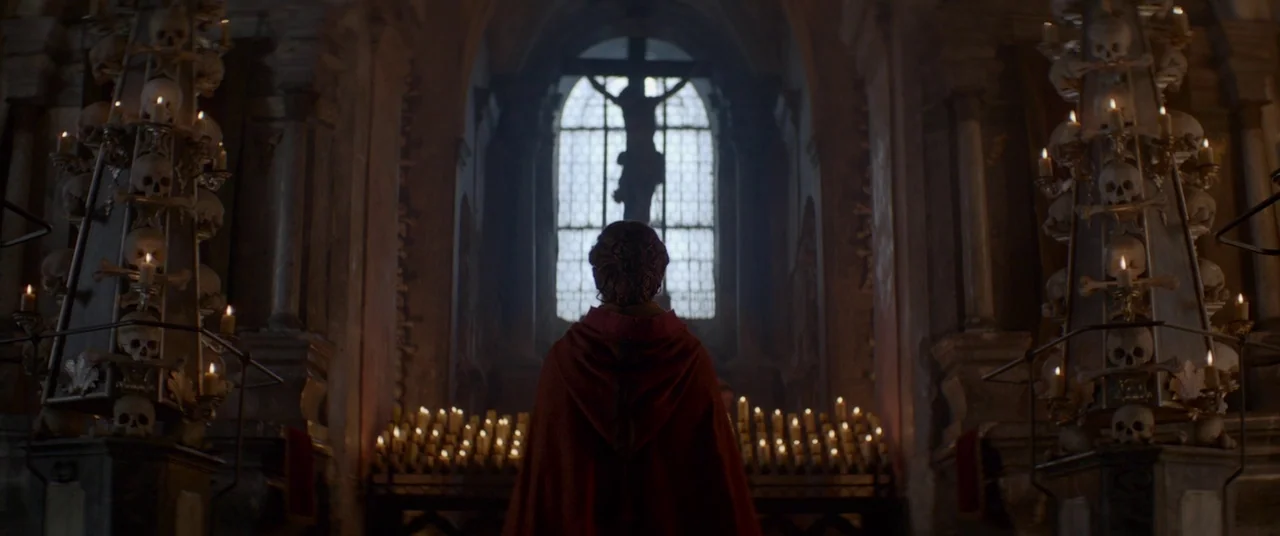 Ophelia (2018) movie still: medium — A dark subterranean space features two tall towers constructed from human skulls and candles. A person wearing…