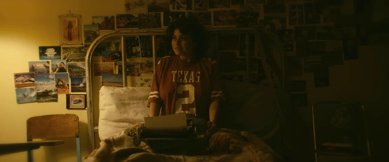 Blaze (2018) movie still: medium — A room with walls covered in numerous vintage travel postcards and photographs. A young woman sits on a bed frame…