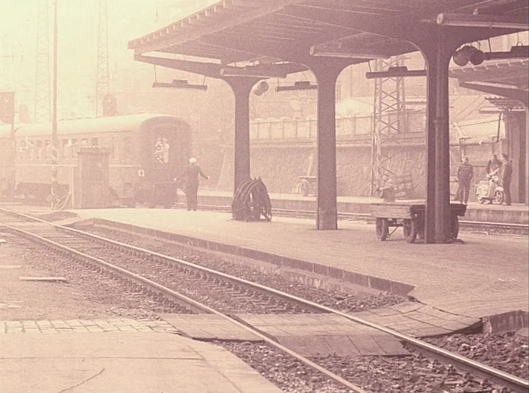 Daisies (1966) movie still: wide — A historic train station platform features a large metal roof structure and tracks. Several people stand near a…