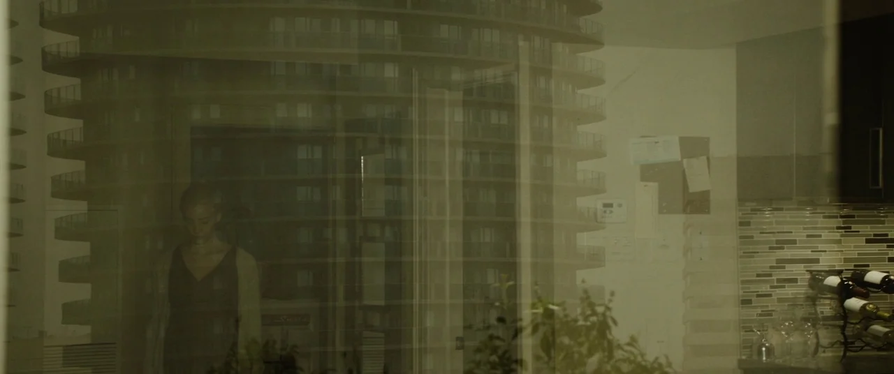 Enemy (2013) movie still: medium — A woman stands inside an apartment behind a glass window reflecting a skyscraper. The glass superimposes the…