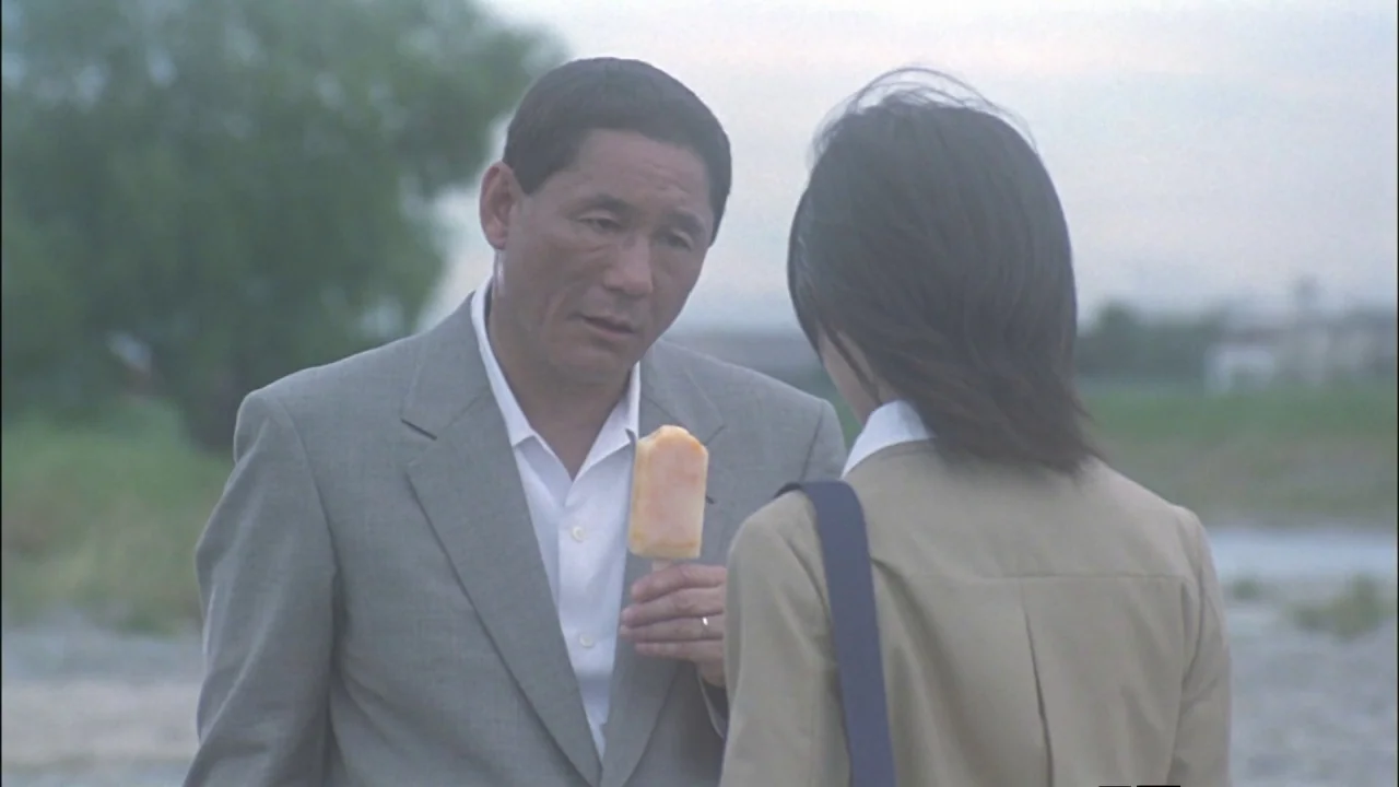 Battle Royale (2000) movie still: medium — An overcast riverbank with sparse vegetation and a blurred landscape in the distance. An older man in a…