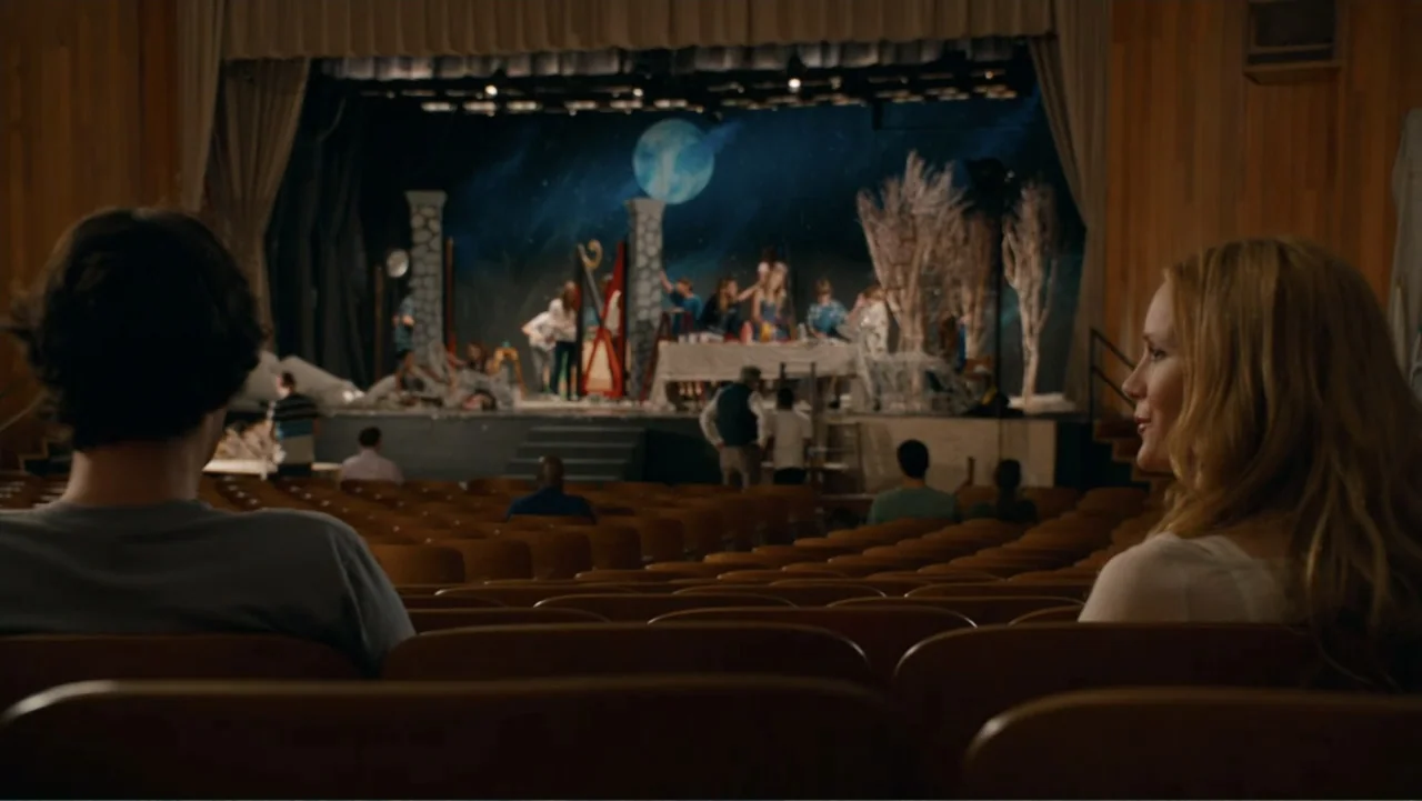 This Is 40 (2012) movie still: medium — A dark auditorium with rows of wooden seats facing a brightly lit stage set. A man and woman sit in the…