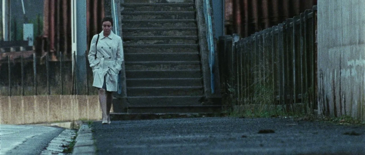 Alléluia (2014) movie still: fullbody — A woman walks down a sidewalk alongside a weathered concrete staircase and fence. The urban environment is…