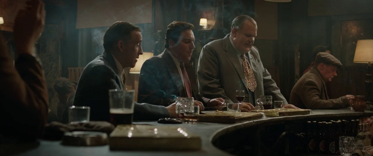 Domino (2005) movie still: medium — A dimly lit bar features a long curved counter with men sitting together. Smoke wisps through the air while the…
