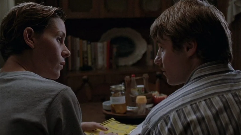 Junebug (2005) movie still: medium — A dark dining room with a wooden hutch filled with plates and books. Two young people sit facing each other at…