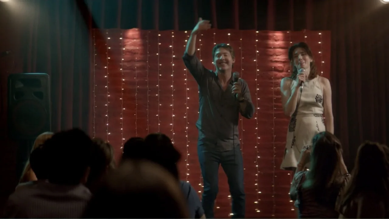 I Married a Dumbass (2016) movie still: medium — A small indoor performance space features a dark red brick wall decorated with fairy lights. A man…