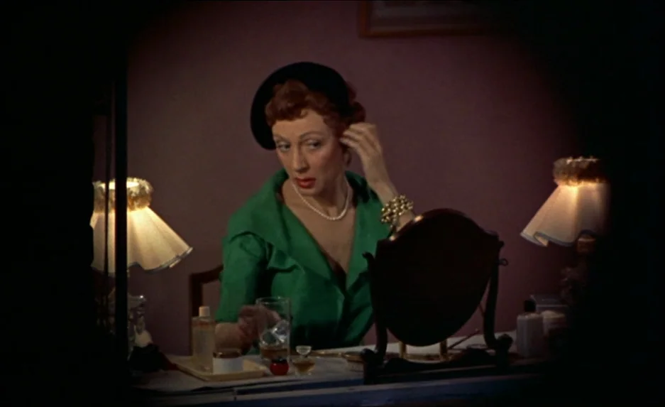 Rear Window (1954) movie still: medium — A dimly lit room features a woman sitting at a vanity table. She adjusts an earring while a glass of liquid…
