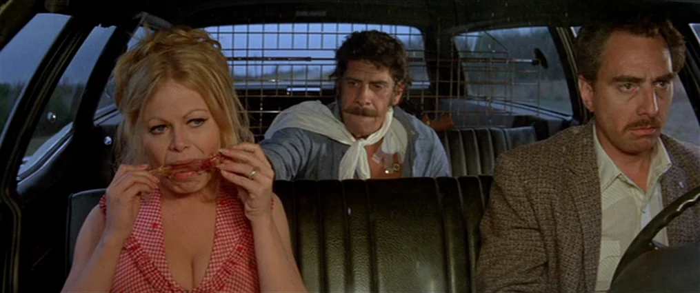 The Getaway (1972) movie still: medium — Three people sit inside a moving car with a metal cage partition separating rows. A blonde woman eats, a…