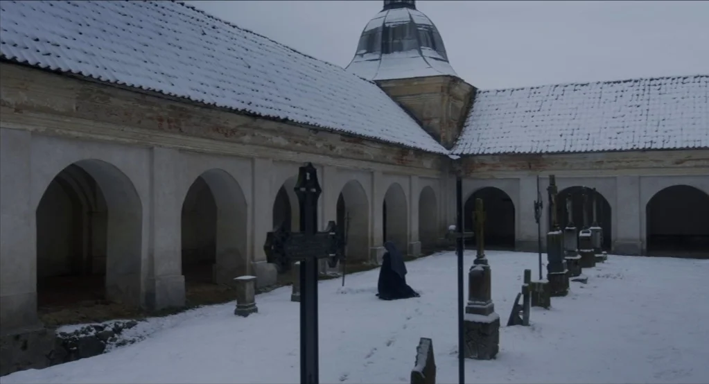 The Innocents (2016) movie still: wide — A stone cloister courtyard with vaulted arches is blanketed in a layer of snow. A person dressed in a dark…
