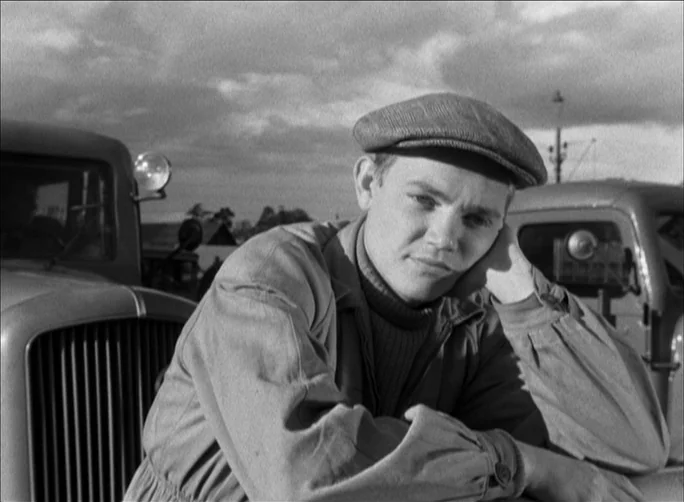 Summer with Monika (1953) movie still: medium — A man stands outdoors positioned between two parked vintage trucks with a cloudy sky. The subject…