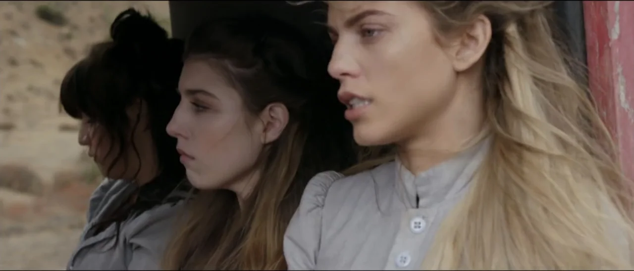 Dead for a Dollar (2022) movie still: medium — Three women sit in profile inside a wooden vehicle with a view of terrain. They gaze off-screen while…