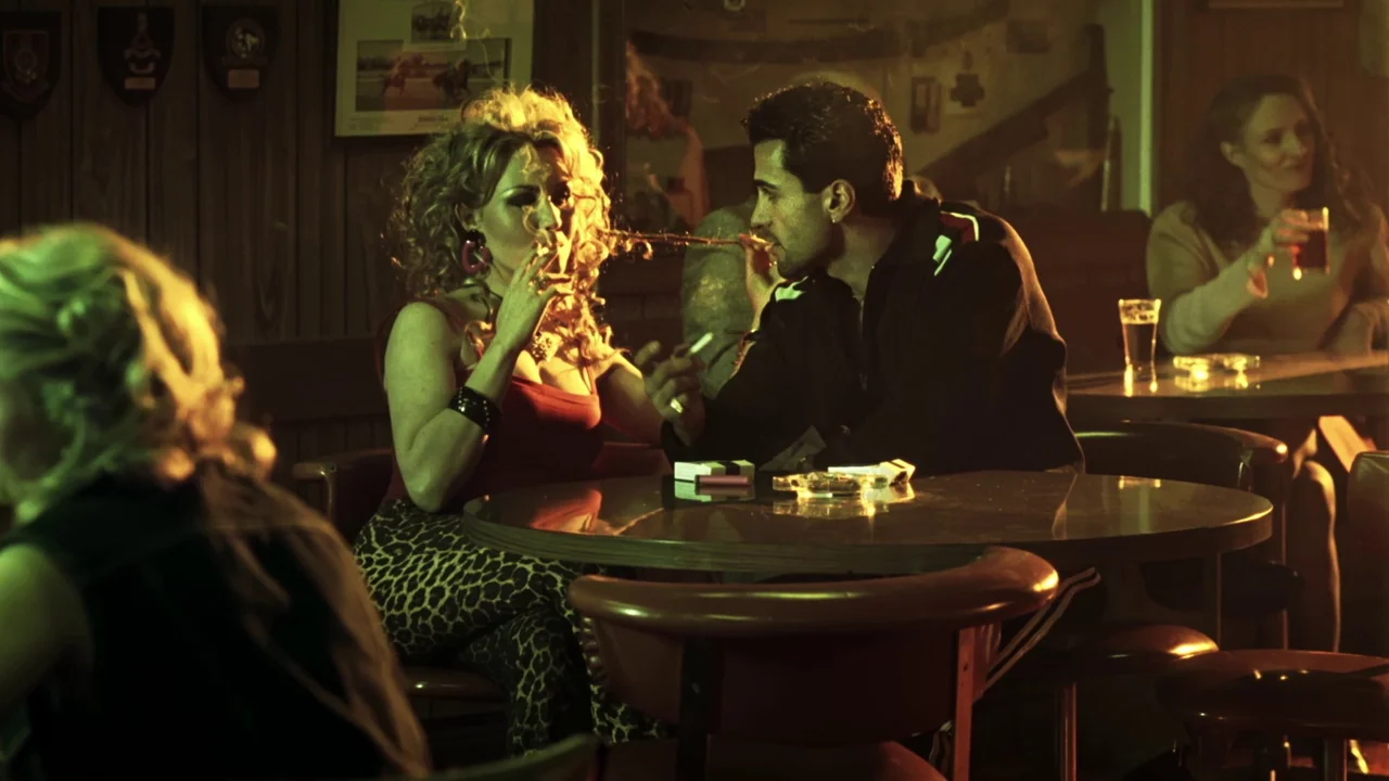 Chopper (2000) movie still: medium — A dimly lit tavern with wood-paneled walls and a circular laminate table. A man and woman sit close together,…