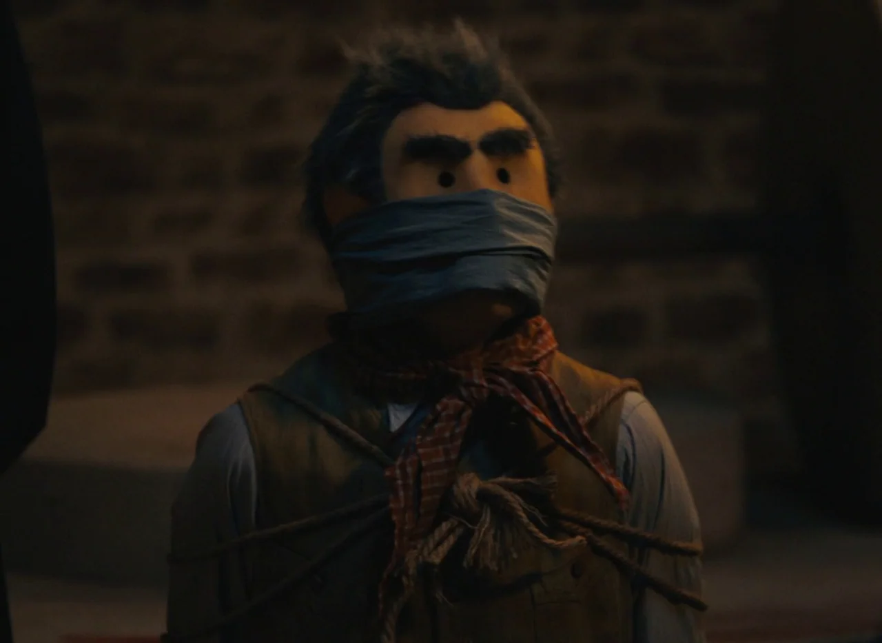 Red Island (2023) movie still: medium — A dimly lit indoor space with a dark brick wall in the background. A puppet tied with ropes stands facing…