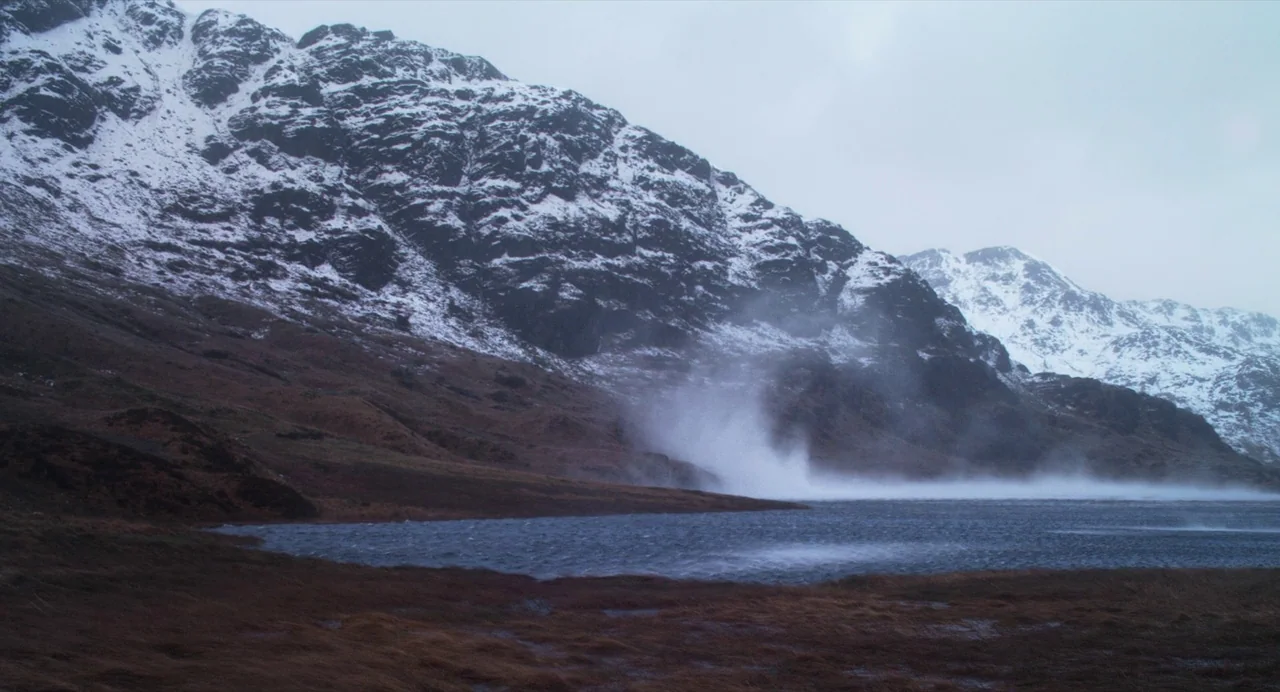 Under The Skin (2013) movie still: wide — A rugged mountain range with snow-dusted slopes rises above a dark, windswept lake. Dense mist swirls…