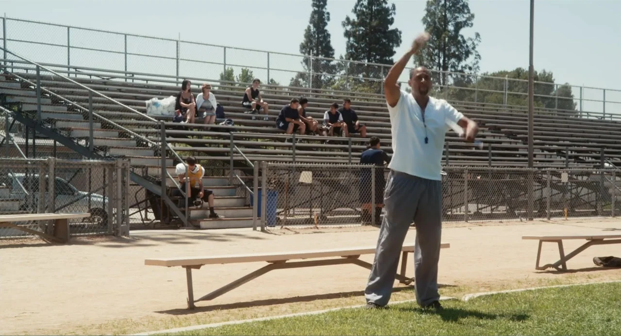 Gates of Darkness (2019) movie still: fullbody — An outdoor sports complex features metal bleachers filled with spectators near a dirt field. A…