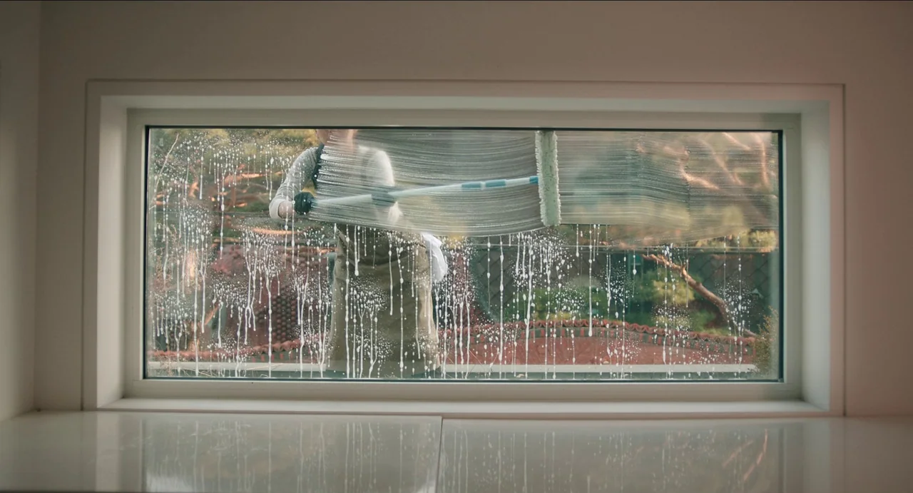 Microhabitat (2017) movie still: medium — A wide, rectangular window displays streaks of soapy water over a blurred outdoor background. A person in…