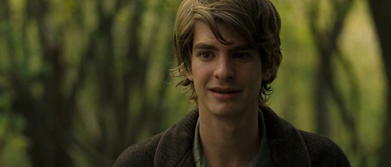 Never Let Me Go (2010) movie still: closeup — A dense forest environment with blurred green foliage forms the backdrop behind a man. The man stands…