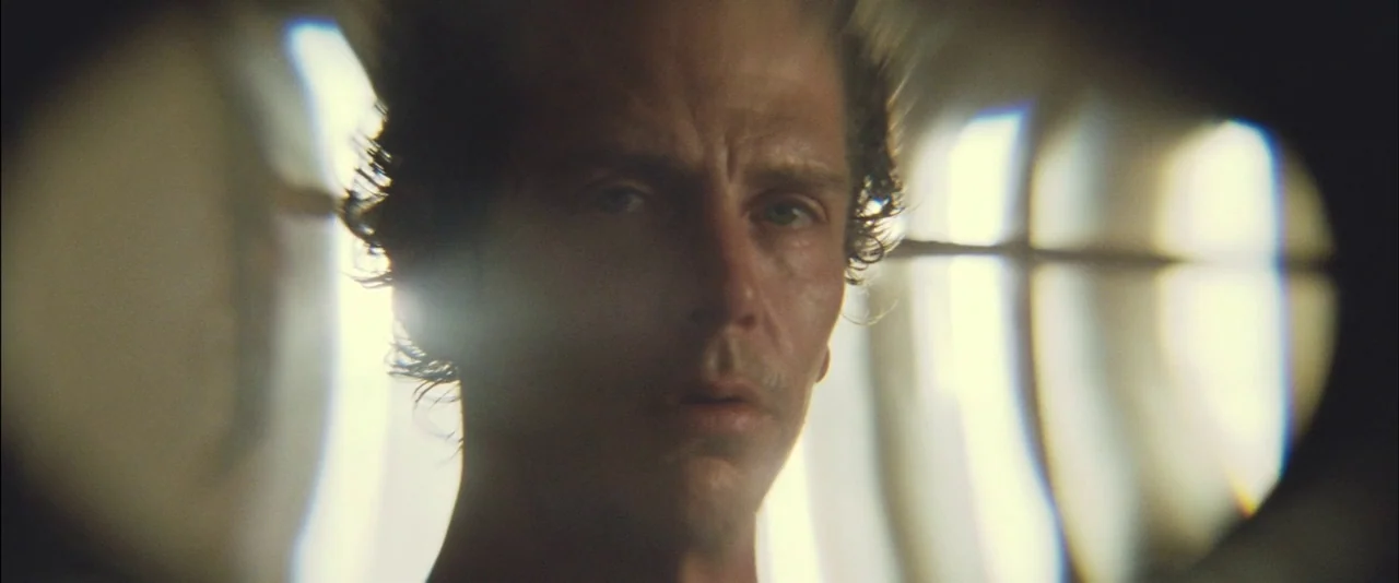 Killing Them Softly (2012) movie still: closeup — A man's face is framed within a narrow, circular peep hole of a door. Bright light spills into the…