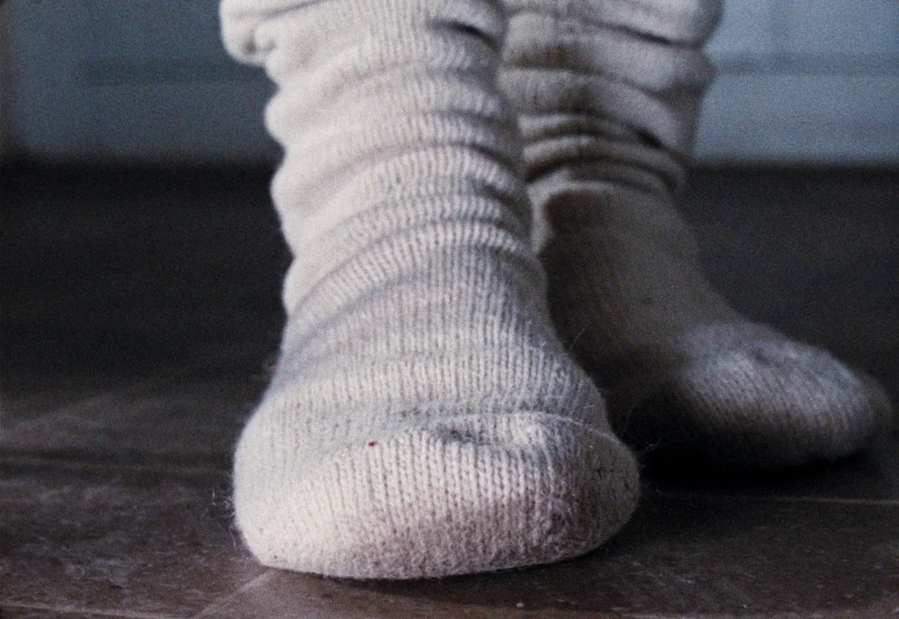 Enys Men (2022) movie still: closeup — A pair of thick, light-colored ribbed socks rests on a textured floor. The soft fabric of the socks is…