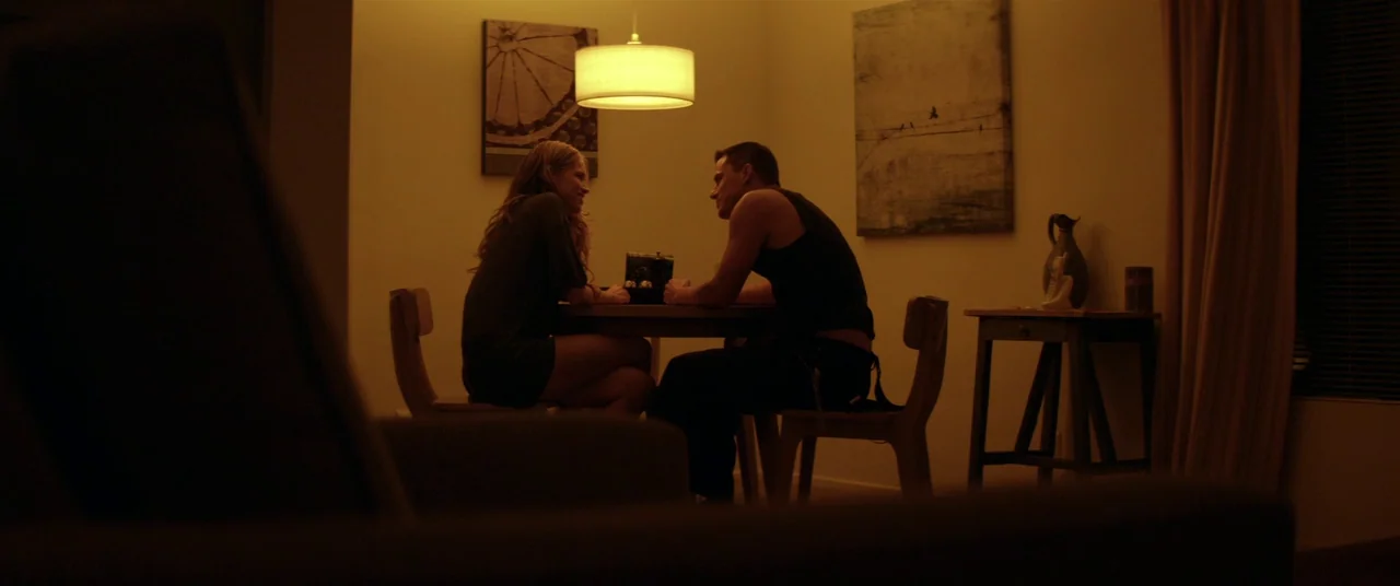 Magic Mike (2012) movie still: wide — A dark, amber-lit room contains a small wooden dining table and two chairs. A man and a woman sit facing each…