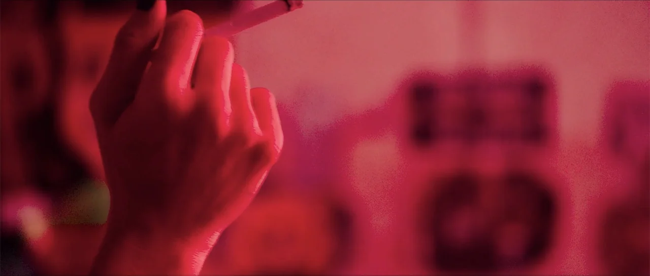 68 Kill (2017) movie still: closeup — A close up shot of a hand holding a cigarette in a dark room. The entire environment is washed in a deep red…