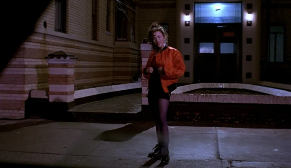 Night on Earth (1991) movie still: fullbody — A young woman stands on a concrete sidewalk at night in front of a building. The brick structure has a…